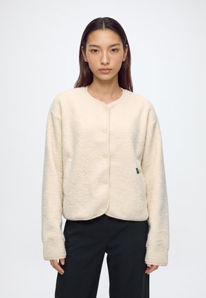 Jeune femme aux cheveux raides et foncés porte une veste polaire crème boutonnée et un pantalon noir, debout devant un fond blanc uni.