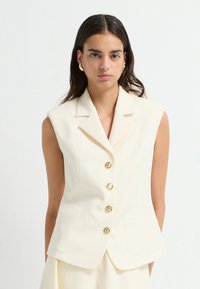 Kremfarvet ærmeløs blazer med fem gyldne knapper, slagkrave og to forlommer, lavet af glat stof med en skræddersyet pasform.