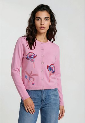Frau mit dunklem, welligem Haar, die eine rosa, geknöpfte Strickjacke mit bunten floralen Stickereien und blaue Jeans vor einem schlichten Hintergrund trägt.