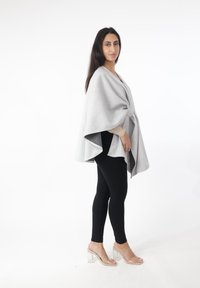 Capa gris tejida con dobladillo asimétrico, atada en la cintura, usada sobre una blusa beige y leggings negros. La modelo lleva sandalias con tacón transparente.