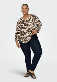 Bluza s svetlo rjavim in kremnim vzorcem, dolgimi rokavi in V-izrezom, v kombinaciji s temnimi kavbojkami in črnimi sandalmi, na skoraj nevtralnem ozadju.