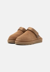 Tan suede slippers with a rounded toe, adjustable strap, and a textured rubber sole. Minimalist design with no visible patterns or accents.
