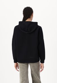 Μαύρη φούτερ μπλούζα με κουκούλα και χαλαρή γραμμή, με ριμπ μανσέτες και στρίφωμα. Η πλάτη εμφανίζει ένα απλό σχέδιο χωρίς διακοσμητικά στοιχεία.