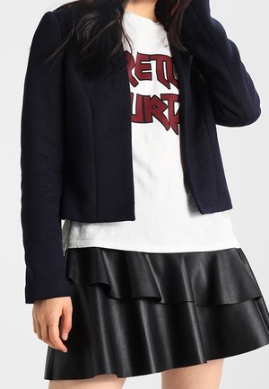 Vrouw draagt een zwart cropped blazer, wit grafisch T-shirt met rode tekst, en een zwarte leren minirok met laagjes.