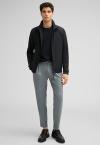 Schwarze Zip-Jacke über einem dunklen T-Shirt, kombiniert mit hellgrauen, maßgeschneiderten Hosen und schwarzen Schnürschuhen. Glatter Stoff und klare Linien.