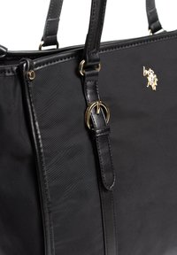 Czarna torba na ramię z gładkiego materiału, skórzanymi akcentami, złotymi elementami metalowymi i detalem z logo. Posiada podwójne skórzane uchwyty oraz przeszycia.