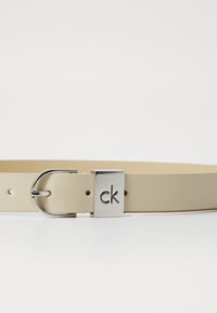 Ceinture en cuir beige avec une boucle en métal argenté arborant un logo "ck" gravé. Texture lisse, avec des trous espacés de manière uniforme pour les ajustements.