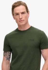 T-shirt verde oliva a maniche corte realizzata in cotone. Presenta un collo a giro e un discreto logo ricamato sul petto. Texture liscia.