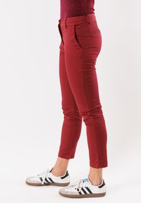 Pantaloni rossi aderenti con tasche laterali, texture liscia e lunghezza cropped. Indossati con sneakers bianche con righe nere.