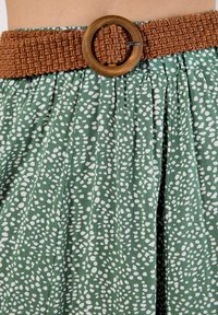 Green fabric skirt with a white speckled pattern, featuring a braided brown belt with a circular wooden buckle. Textured and pleated design.