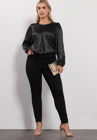 Top de satén negro de manga larga con pliegues, combinado con pantalones ajustados negros. Lleva un clutch dorado y sandalias de tacón dorado.