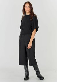 Melnā crop top ar īsām piedurknēm, apvienots ar platām melnām biksēm un melnām ādas zābakām līdz ceļiem. Gluda tekstūra, minimāls dizains.