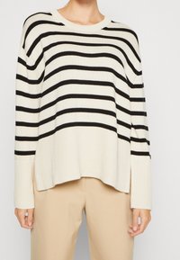 Creme- und schwarz gestreifter Strickpullover mit gerippter Struktur, lockerer Passform und Seitenschlitzen, kombiniert mit beigen Hosen.