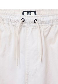 Helle beige Shorts mit einem elastischen Bund und Kordelzug, schwarzen Ösen und einem Etikett mit einem Logo. Glattes Material mit minimaler Textur.