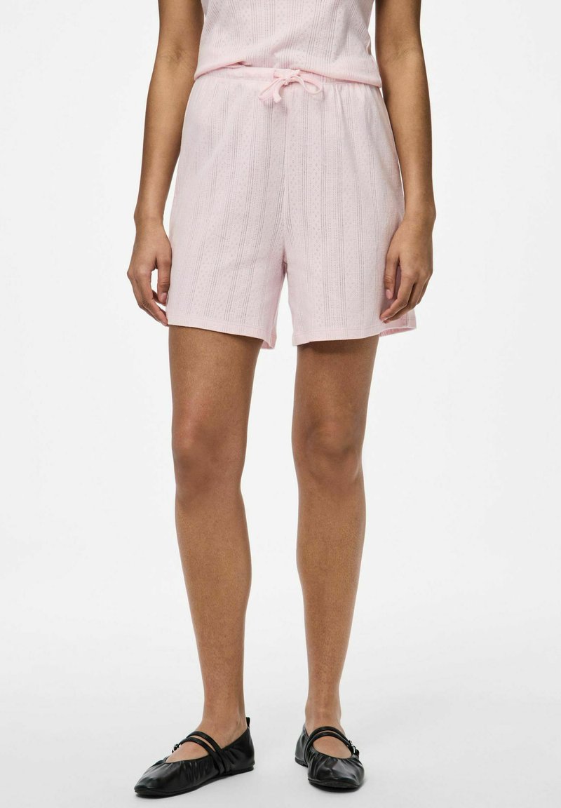 Pantalones cortos de color rosa claro con un patrón texturizado, una cinturilla elástica con cordón y un ajuste relajado. Llevados con zapatos planos negros.