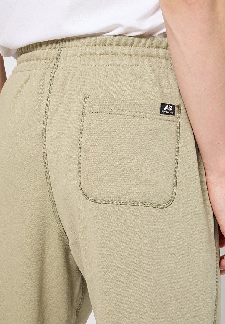 Pantalons de jogging beige New Balance avec taille élastique et poche arrière portés par une personne en chemise blanche, vue recadrée de la taille jusqu'en bas.