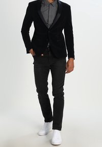 Czarna aksamitna marynarka z satynowymi klapami, wzorzysta koszula, czarne dżinsy i białe sneakersy. Posiada dopasowany krój i klasyczną sylwetkę.