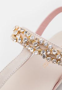 Beige mockasinsandal med rosa rem, med två rader av klara och gyllene strass längs ovansidan. Slät, texturerad yta.