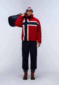 Giacca di pile rossa con un design incrociato bianco e navy, pantaloni navy, stivali rossi con lacci e un berretto azzurro chiaro con la bandiera norvegese.