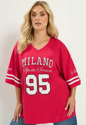 Lyshåret kvinde iført oversized rød V-hals trøje med hvid tekst "Milano Sports Season 95" og stribede ærmer, kombineret med blå jeans.