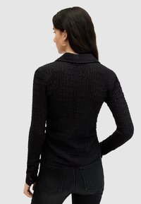 Top lungo nera a maniche lunghe con colletto, caratterizzata da un motivo ondulato e dettagli con bottoni sui polsini, abbinata a jeans scuri.