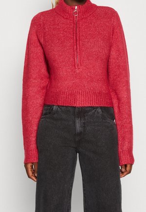 Personne portant un pull en tricot rouge avec une fermeture éclair à l'avant et un col montant, associé à un pantalon en jean noir taille haute.