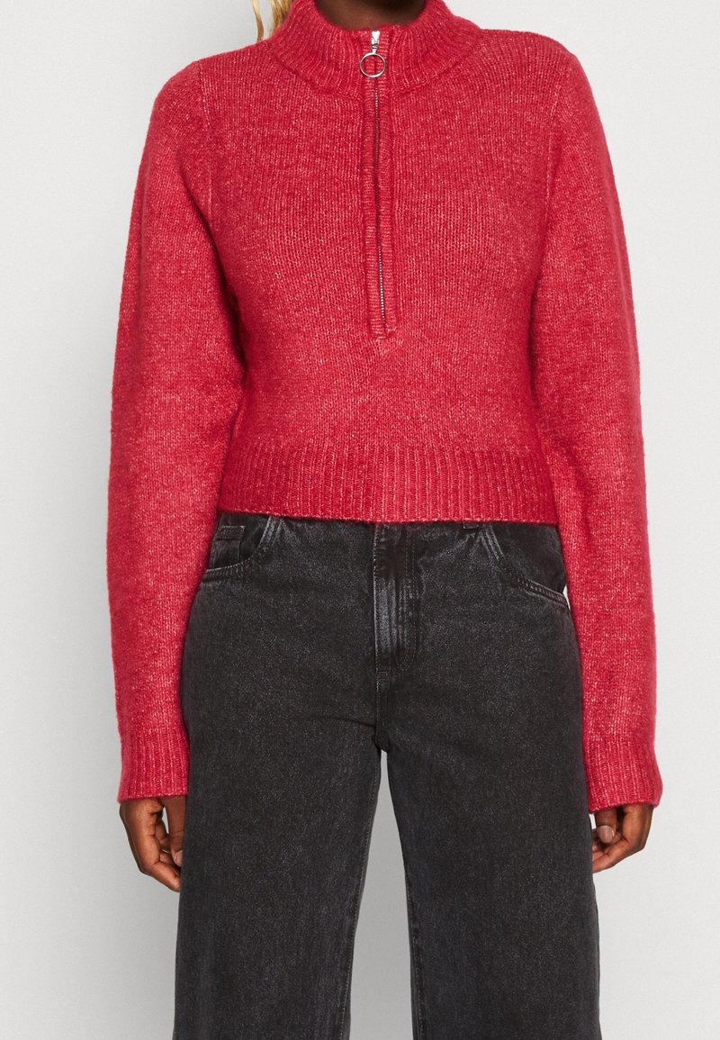 Personne portant un pull en tricot rouge avec une fermeture éclair à l'avant et un col montant, associé à un pantalon en jean noir taille haute.