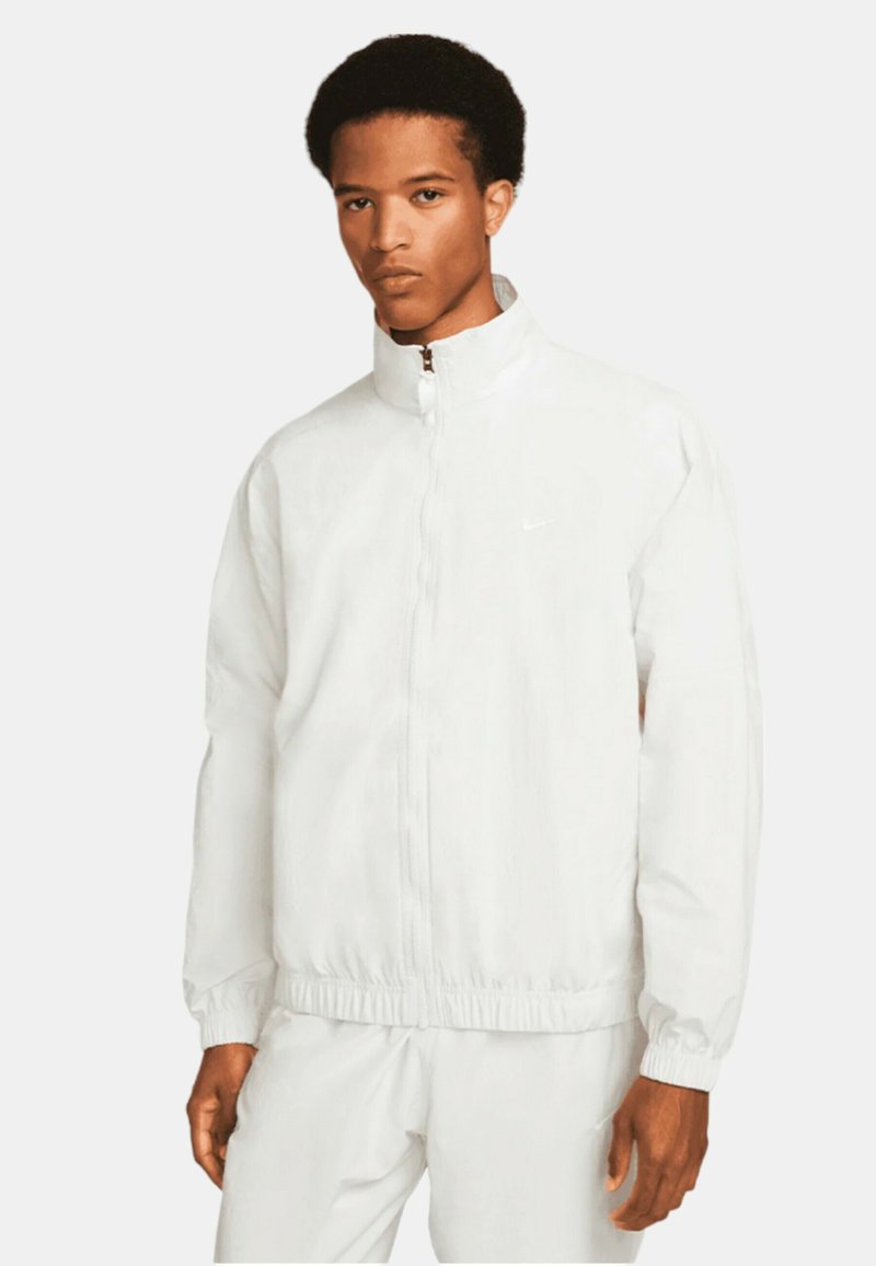 Veste blanche zippée avec col haut et poignets élastiqués. Présente un logo Nike discret sur la poitrine. Texture lisse, matériau léger.