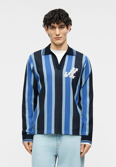 Jeune homme aux cheveux bouclés portant une chemise à manches longues rayée verticalement bleu et noir avec un logo blanc sur la poitrine et un jean bleu clair.