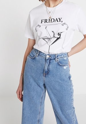 Kobieta nosząca białą koszulkę z napisem „FRIDAY,” i grafiką kieliszka do martini, w zestawieniu z jasnoniebieskimi jeansami z wysokim stanem i warstwowymi naszyjnikami.