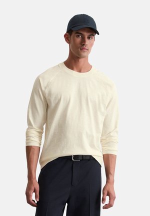 Chemise crème à manches longues avec des manches raglan, assortie à un pantalon marine et une casquette sombre. Le tissu semble doux avec un design simple.