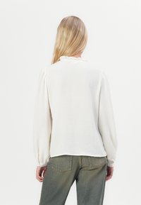 Biała bluzka z długim rękawem, z falbanistym kołnierzem i teksturowanym materiałem, w połączeniu z zielonymi spodniami o wysokim stanie. Miękki, lekki materiał.