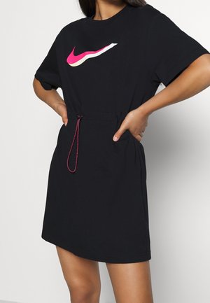 Femme portant une robe noire à manches courtes avec un logo swoosh rose et blanc et un cordon rose à la taille.