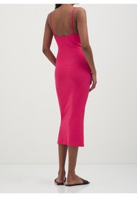 Femme portant une robe midi sans manches rose vif avec des fines bretelles, debout pieds nus sur un sol blanc devant un fond clair uni.