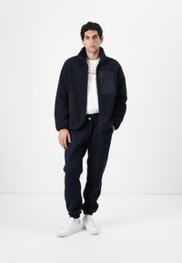 Jachetă din fleece bleumarin cu guler înalt și buzunar frontal, asociată cu pantaloni de trening bleumarin și adidași albi. Design simplu și casual.