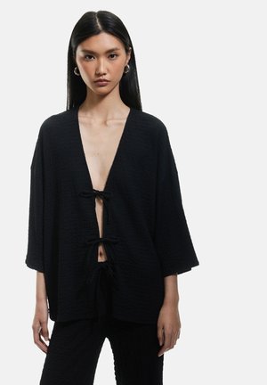Zwart textuur kimono-stijl top met een diepe V-hals, een gestrikte voorkant en brede mouwen. De stof heeft een zachte, gekreukte uitstraling.
