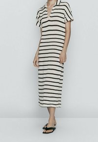 Donna che indossa un vestito midi a maniche corte e scollo a V di colore crema con strisce orizzontali nere e sandali aperti neri senza chiusura.