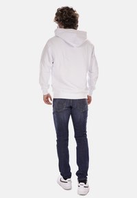 Felpa bianca con cappuccio e tasca a marsupio, abbinata a jeans scuri e sneakers bianche. La felpa presenta un cappuccio regolabile con lacci.