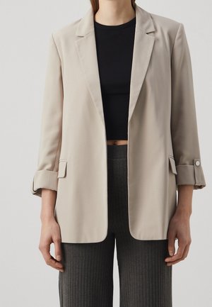 Femme portant un blazer beige avec les manches retroussées sur un top court noir et un pantalon côtelé foncé.