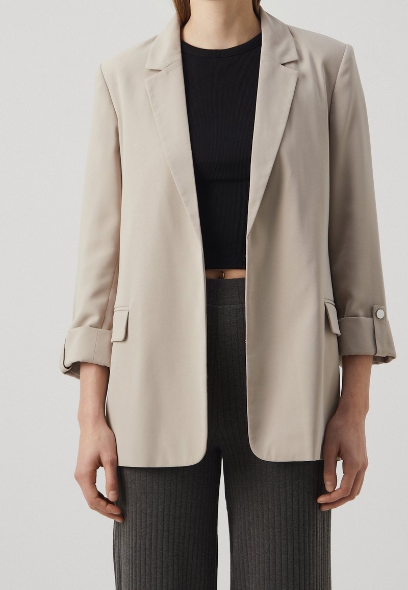 Femme portant un blazer beige avec les manches retroussées sur un top court noir et un pantalon côtelé foncé.