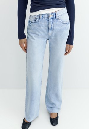 Person trägt hellblaue Jeans mit weitem Bein, dunkelblauen Langarmtop und schwarze Ballerinas, steht vor einem schlichten weißen Hintergrund.