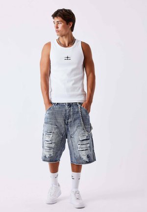 Jeune homme portant un débardeur blanc et un short en jean déchiré avec une chaîne, debout les mains dans les poches, regardant sur le côté.