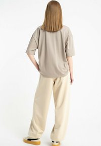 Osoba z prostymi, sięgającymi ramion włosami, ubraną w luźny beżowy t-shirt, kremowe spodnie o szerokich nogawkach oraz beżowe sneakersy na gumowej podeszwie, stojąca z jedną ręką na biodrze.