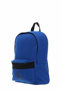 Sac à dos en tissu bleu avec un dessus arrondi, des accents noirs, une poche frontale zippée et un détail de logo en or. Design simple et fonctionnel.