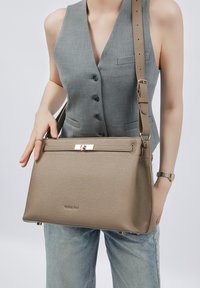 Beige Lederhandtasche mit strukturierter Oberfläche, goldfarbenen Beschlägen und einem Vorderklappenverschluss. Verstellbarer Riemen und dezent geprägtes Logo an der Unterseite.