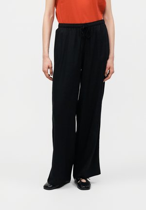 Persoană purtând pantaloni negri largi cu șnur, top portocaliu fără mâneci și balerini negri, stând pe un fundal simplu.