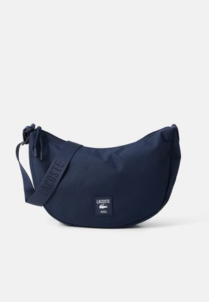 Mørkeblå halvmåneformet crossover-veske med justerbar stropp, glidelås-lukking og Lacoste Paris-logo på fronten.