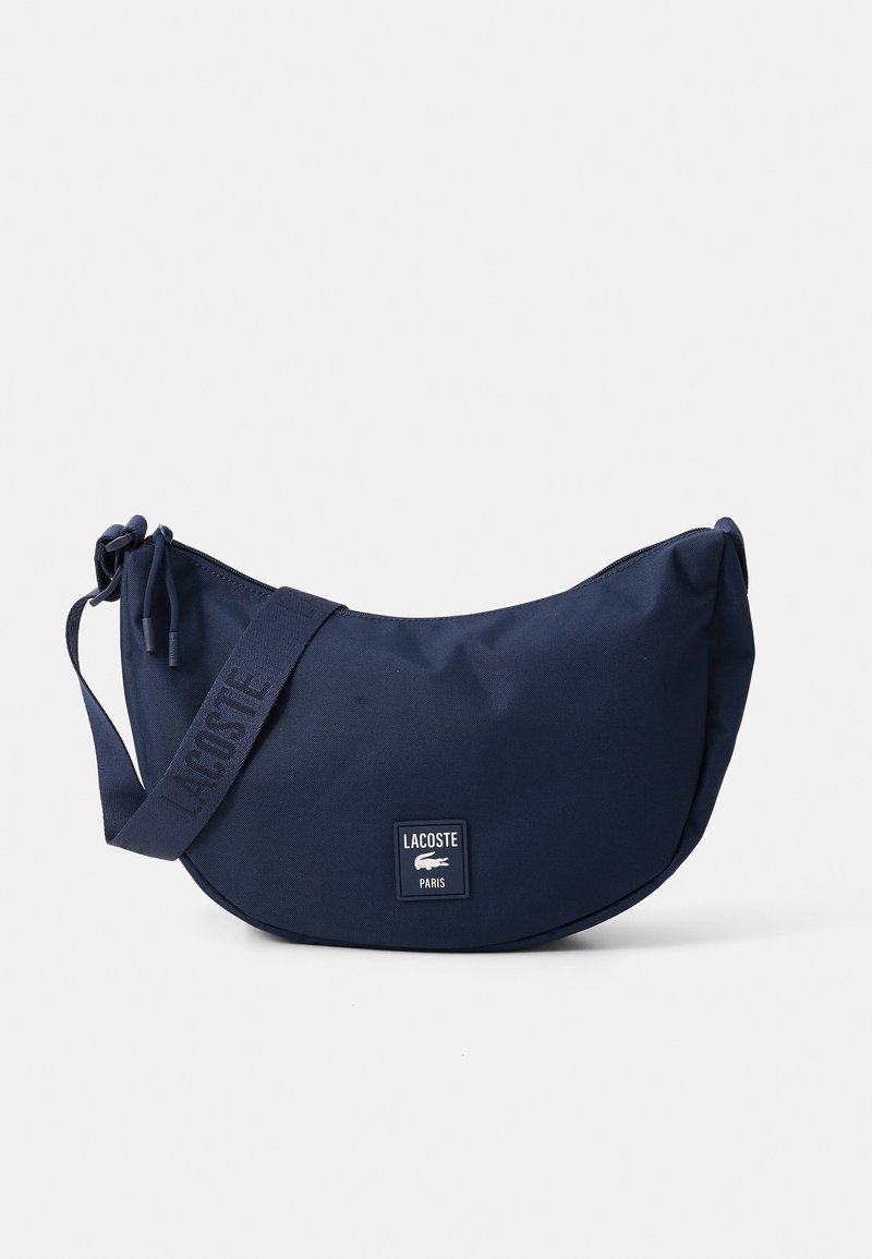 Mørkeblå halvmåneformet crossover-veske med justerbar stropp, glidelås-lukking og Lacoste Paris-logo på fronten.