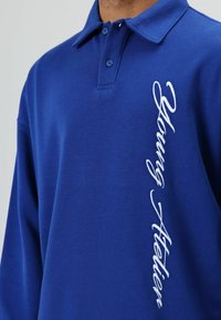 Blauw poloshirt van katoen met een kraag en twee knopen. Witte borduurwerk met de tekst "Young Atelier" verticaal aan de linkerkant.