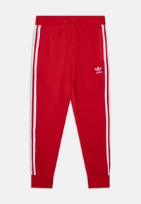 Pantalones de chándal rojos con franjas laterales blancas, cintura elástica con cordón y un logo de Adidas impreso en blanco en el muslo izquierdo.